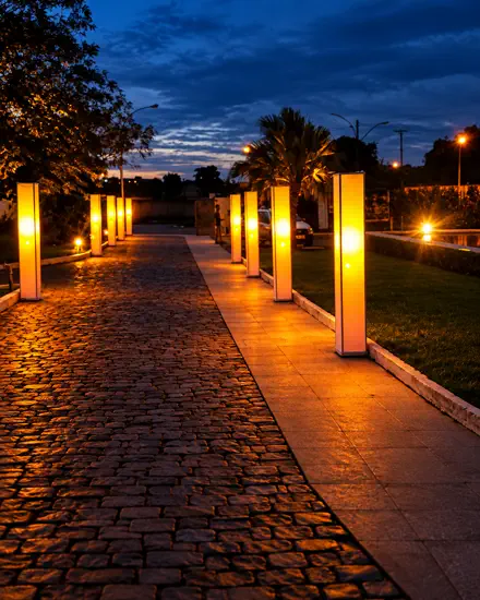 Caminho de entrada com duas fileiras de totens de luz âmbar acesos, refletidos no piso de pedra ao anoitecer