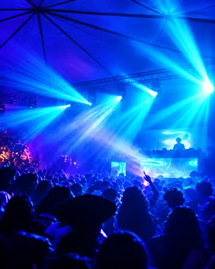 Público sob a tenda atravessado por feixes azuis, com a cabine de DJ e os painéis de LED acesos ao fundo