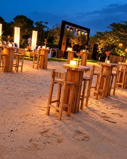 Lounge na areia ao entardecer, com banquetas de madeira, totens de luz e palco ao fundo